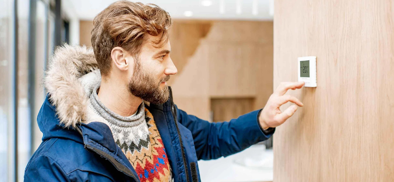 A bearded man in a blue winter jacket adjusts a modern thermostat on a wooden wall in a bright room.